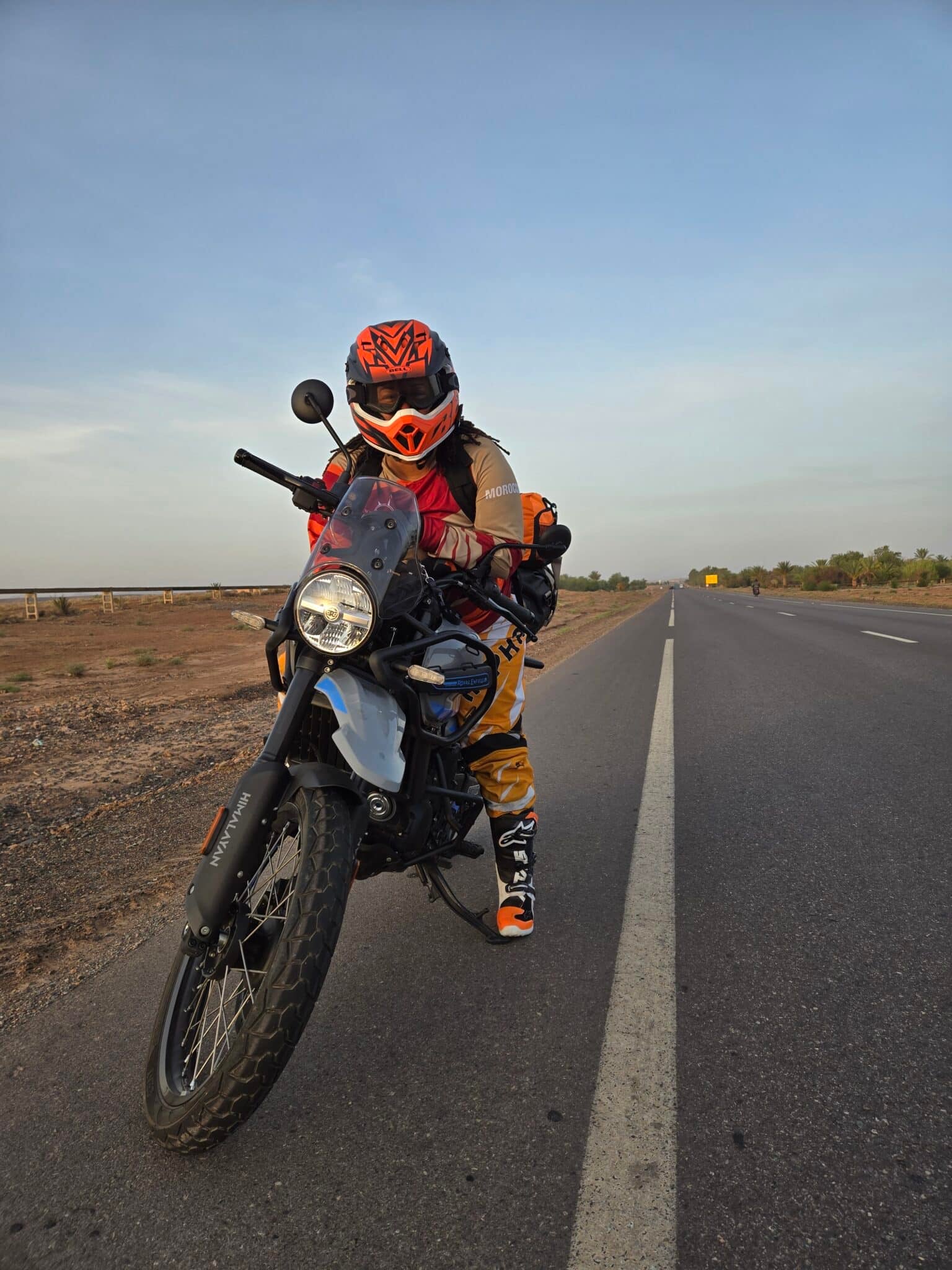 Motorcyclist in orange and black gear leaning on a black dual-sport motorcycle on an empty road with clear sky.