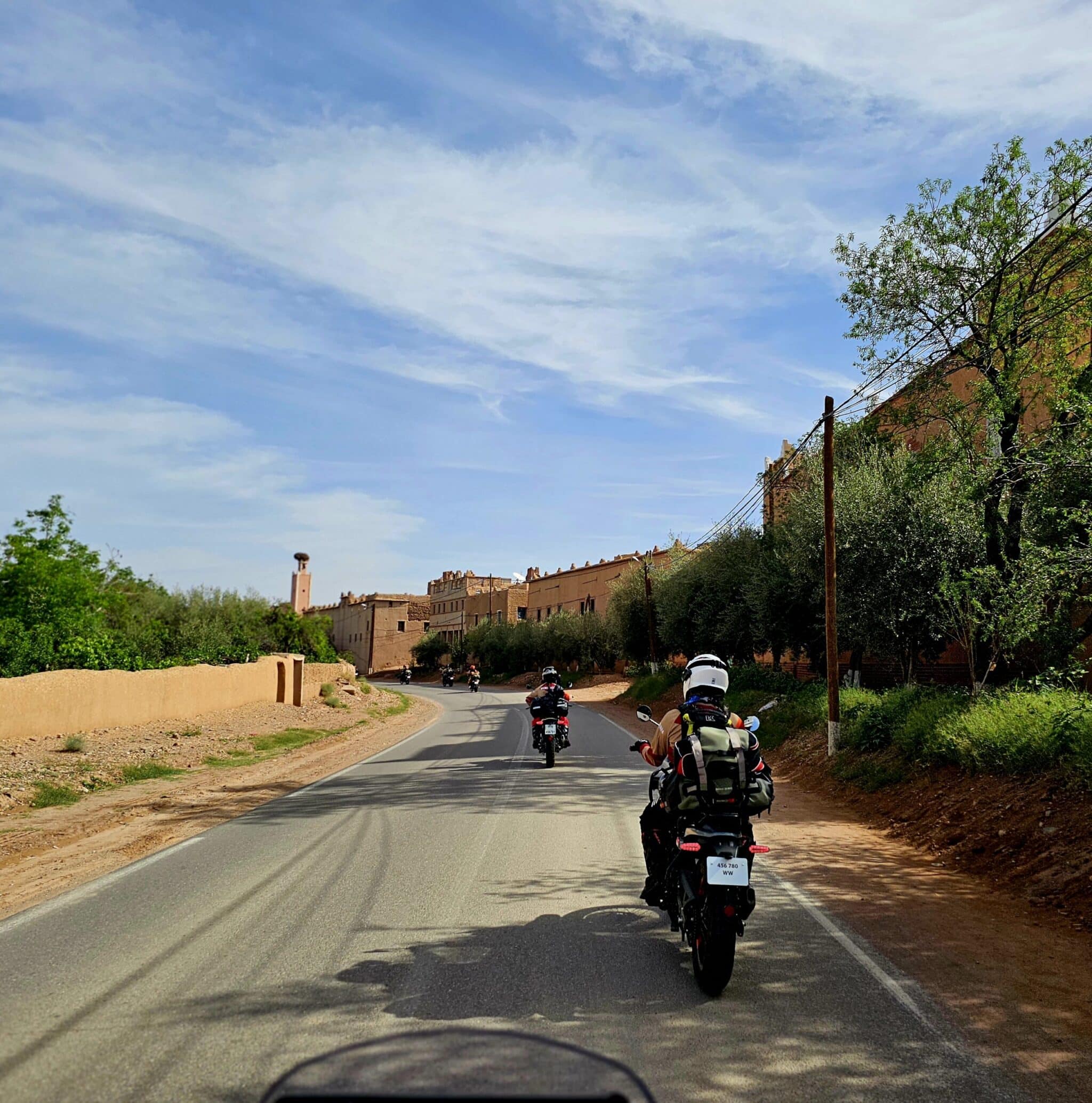 Motorcyclists riding on a scenic road surrounded by lush greenery and rustic buildings under a blue sky with clouds, ideal for adventure travel and exploration.