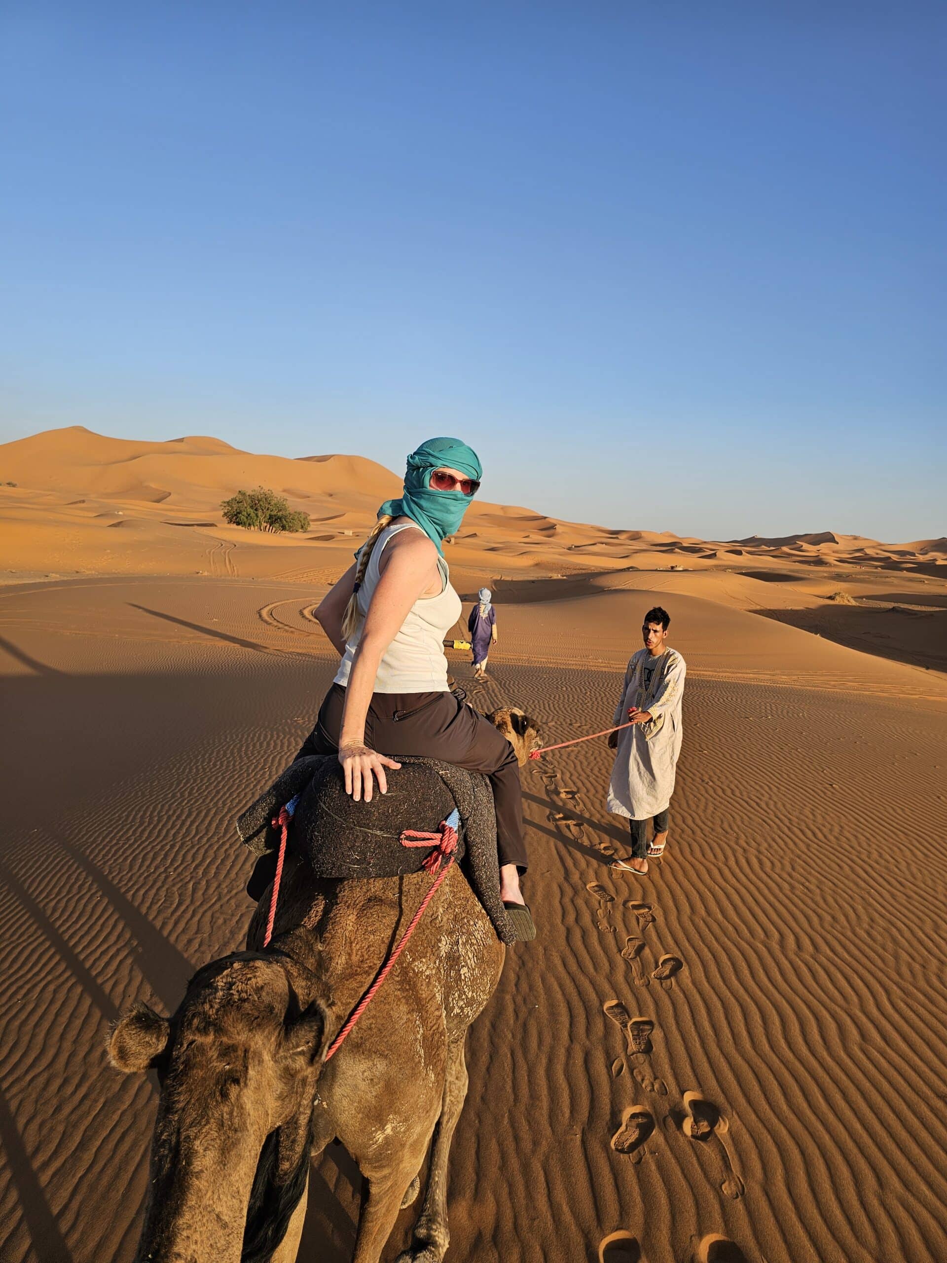 A traveler wearing a teal scarf rides a camel in a desert landscape, with sand dunes in the background and a local guide walking alongside. The scene captures the adventure of camel trekking in a warm, sunny environment.