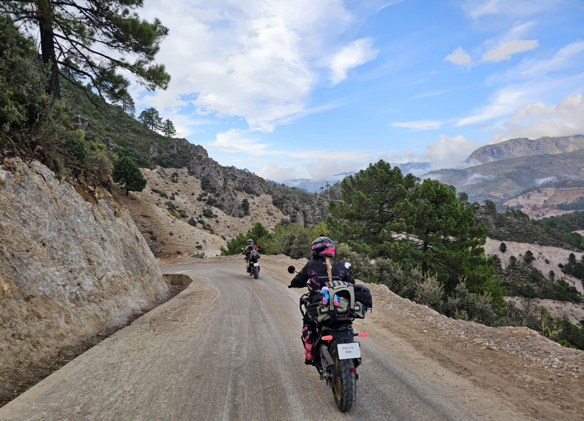 Motorcyclists riding on a scenic dirt road surrounded by mountains and trees, under a partly cloudy sky.