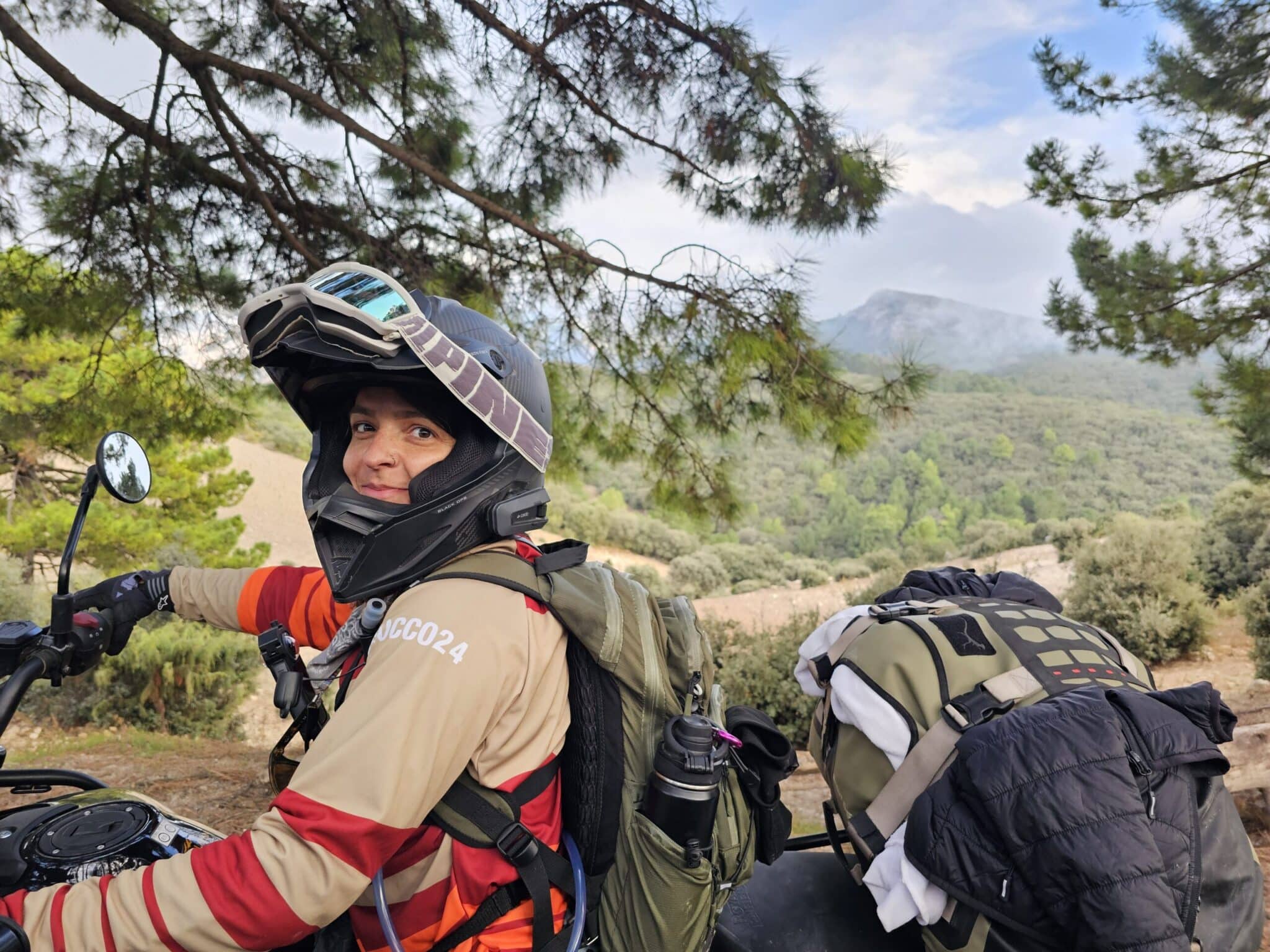 A motorcyclist wearing a helmet and protective gear smiles while sitting on an off-road motorcycle, surrounded by lush greenery and mountains in the background, showcasing an adventurous outdoor lifestyle.