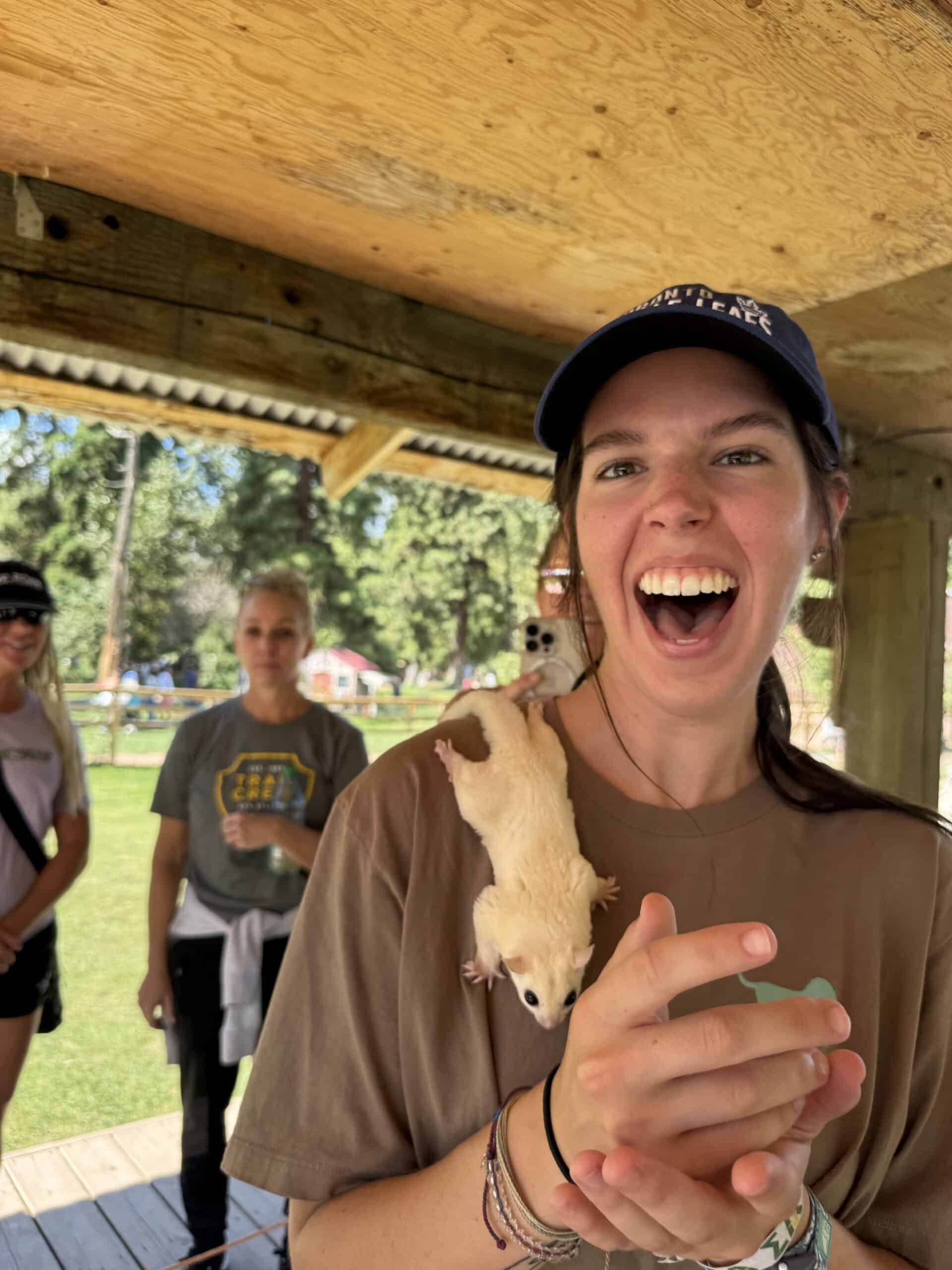 Young woman smiling with a small cream-colored sugar glider on her shoulder, outdoors under a wooden shelter with two women in the background.