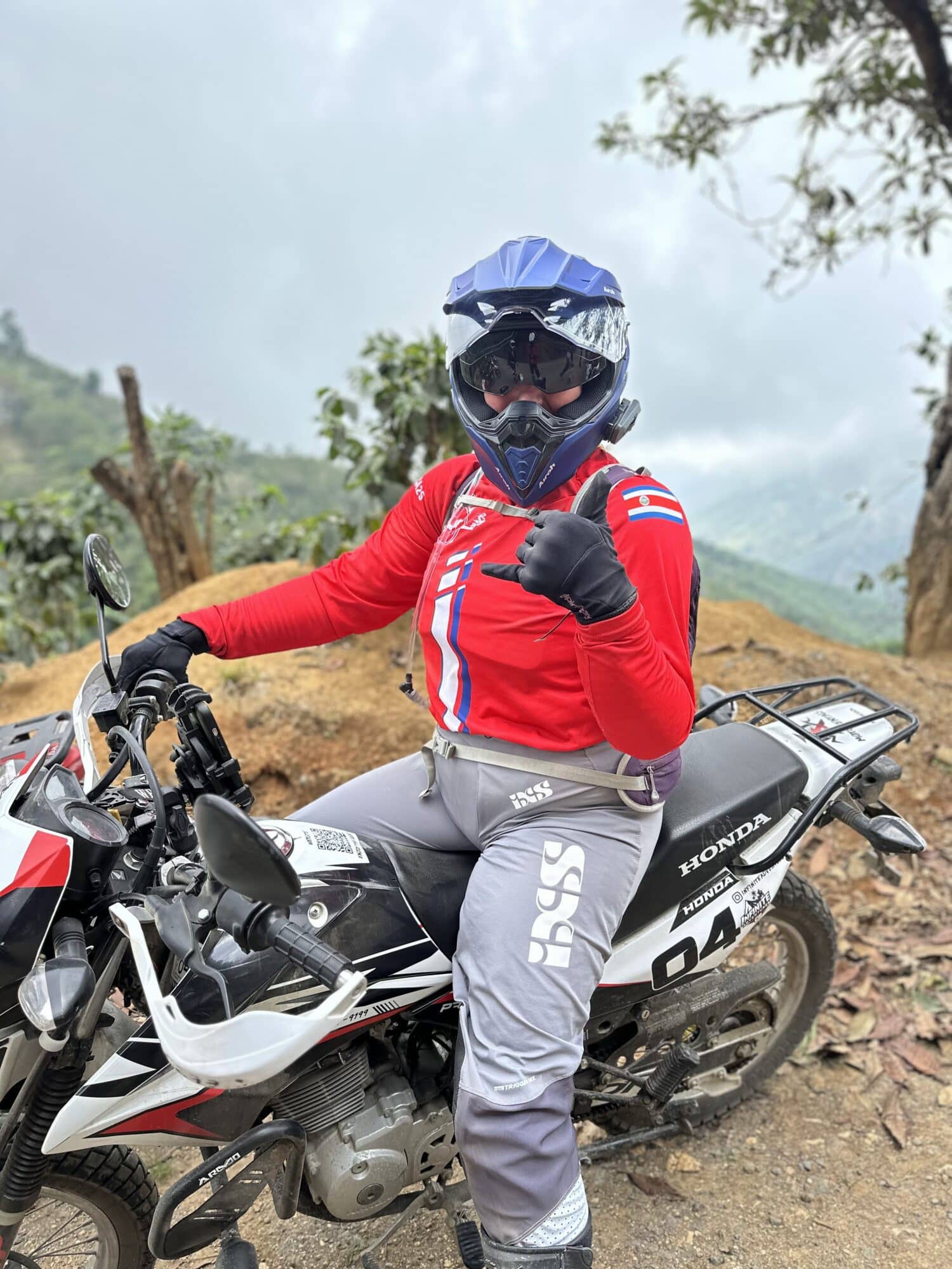 Off-road motorcycle rider giving a thumbs up while wearing protective gear, including a helmet and red jacket, in a scenic mountainous landscape.