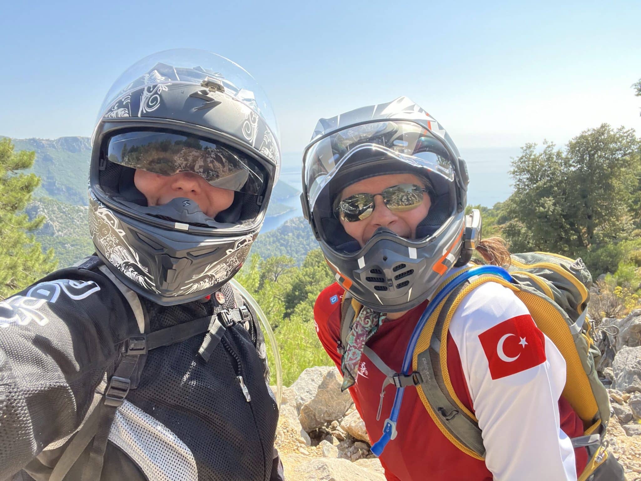 Two motorcyclists wearing helmets and protective gear pose for a selfie on a scenic mountain overlook, showcasing a beautiful landscape in the background. One rider has a Turkish flag on their backpack, indicating a connection to Turkey.
