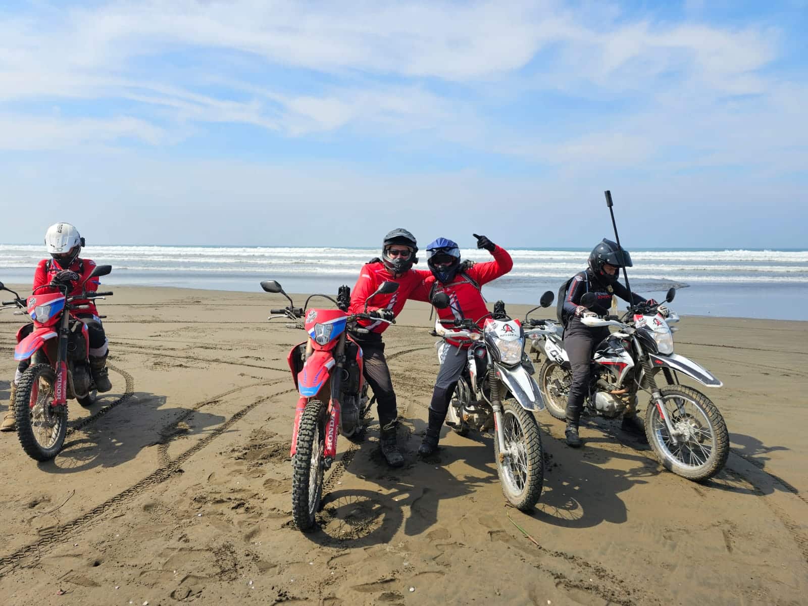 Group of motorcyclists in red jackets posing on a sandy beach with ocean waves in the background, showcasing an adventurous day of off-road biking.