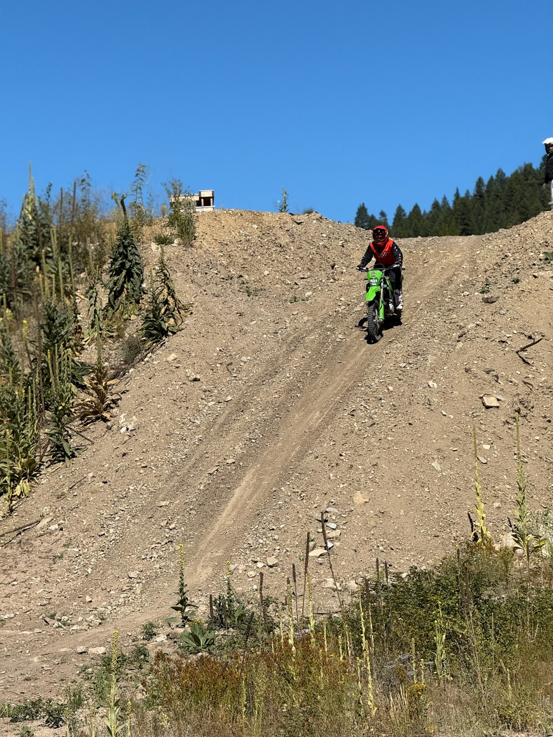 Person riding a green dirt bike downhill on a rocky dirt trail with clear blue sky and greenery in the background.