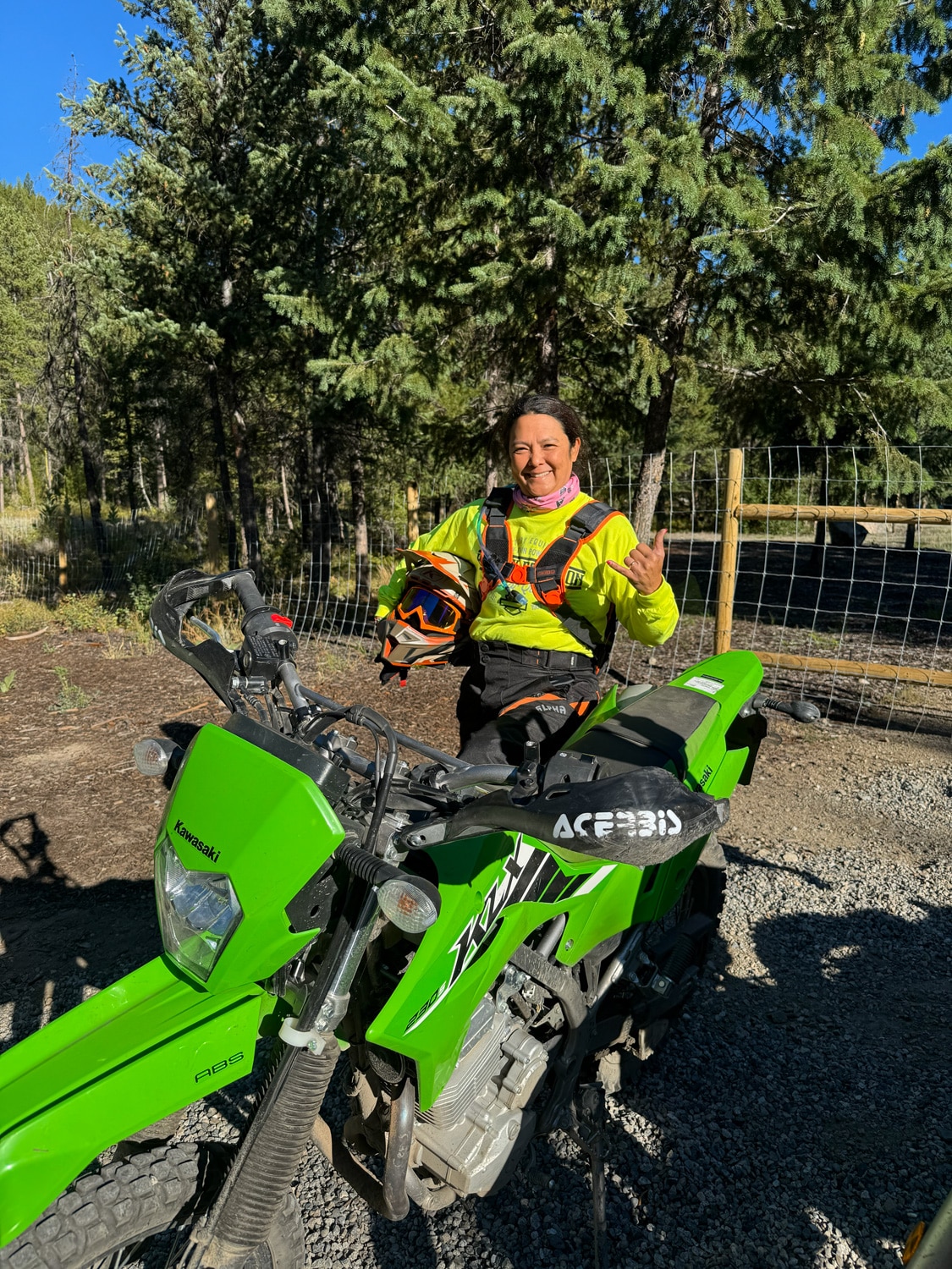 Woman in bright yellow riding gear posing with green Kawasaki dirt bike in forested outdoor area.
