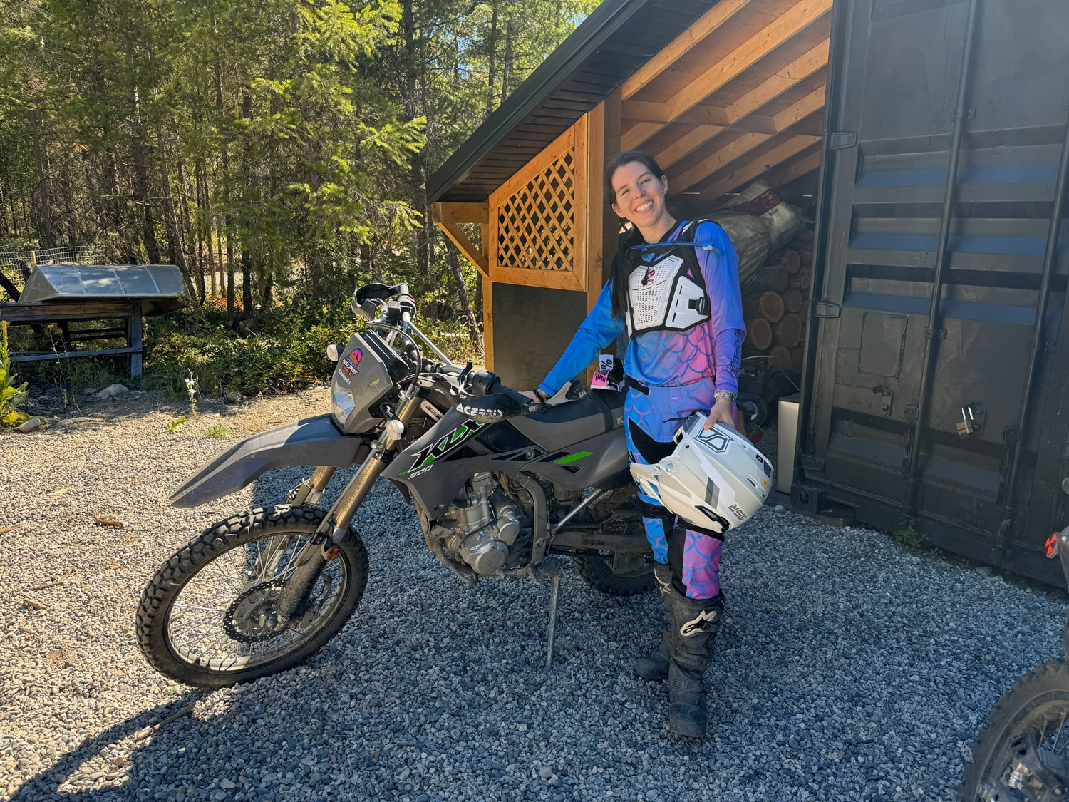 Woman in blue and pink motocross gear standing next to a black and green Kawasaki dirt bike on gravel near a wooden shed and trees.