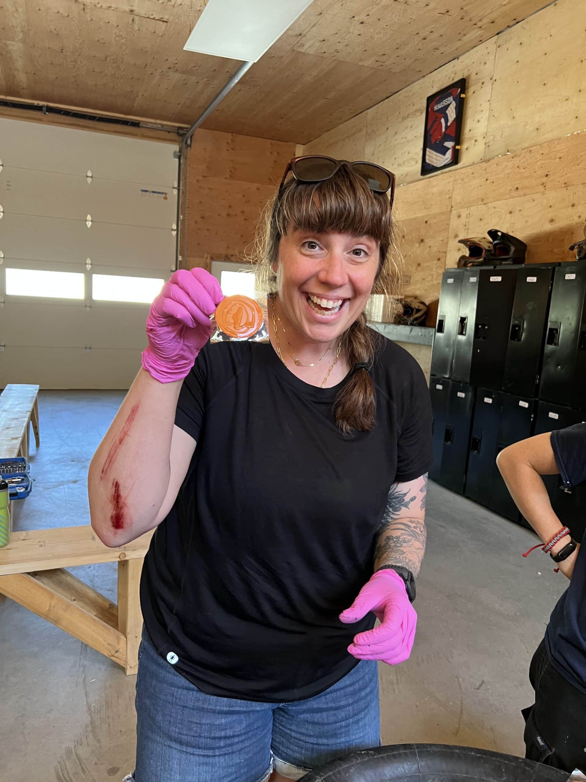 Woman wearing pink gloves and a black shirt showing a packaged orange item, with a scrape on her arm, inside a garage or workshop with lockers in the background.