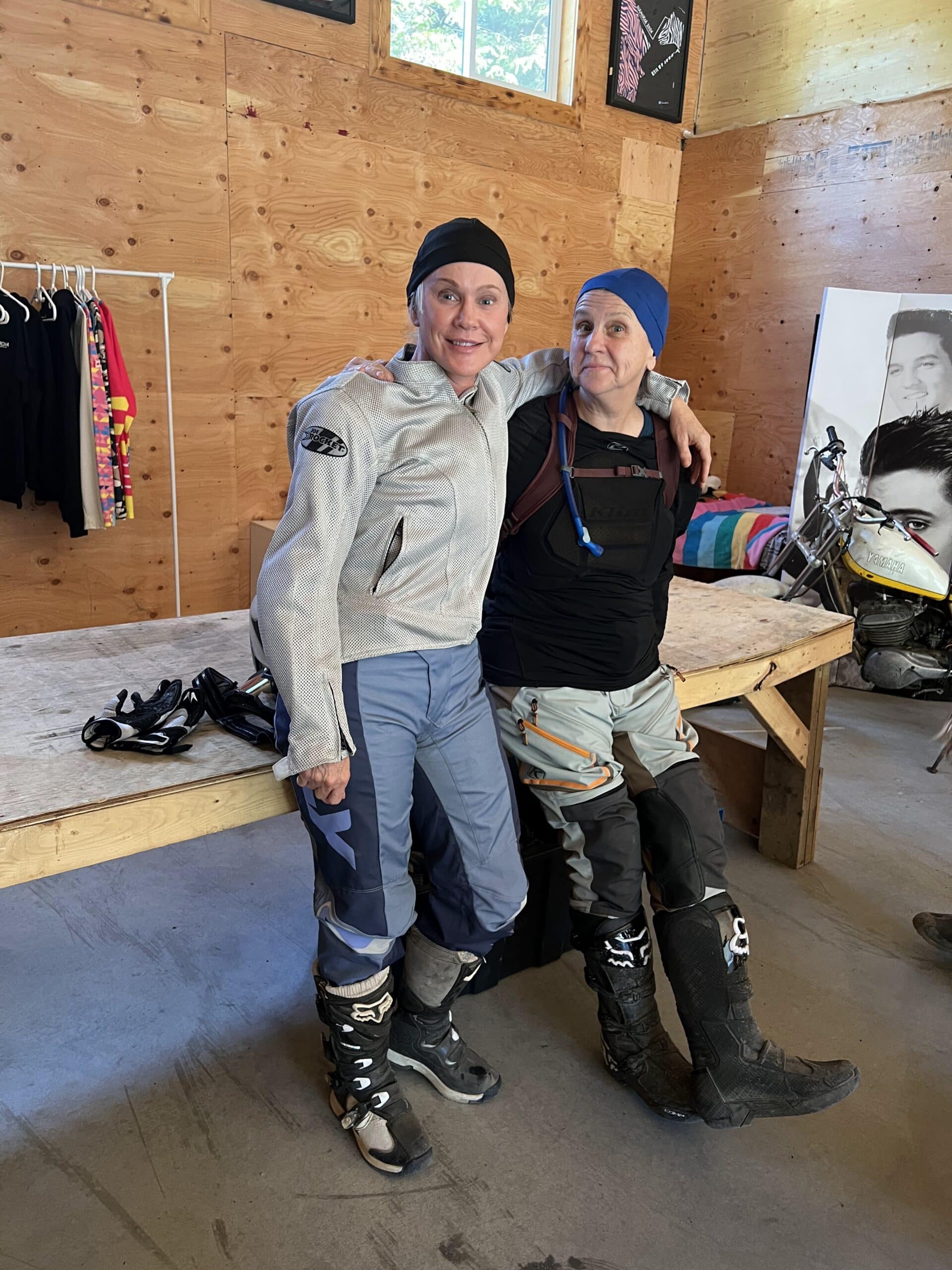 Two women in motocross gear posing inside a wooden garage with helmets and gloves on a table behind them.