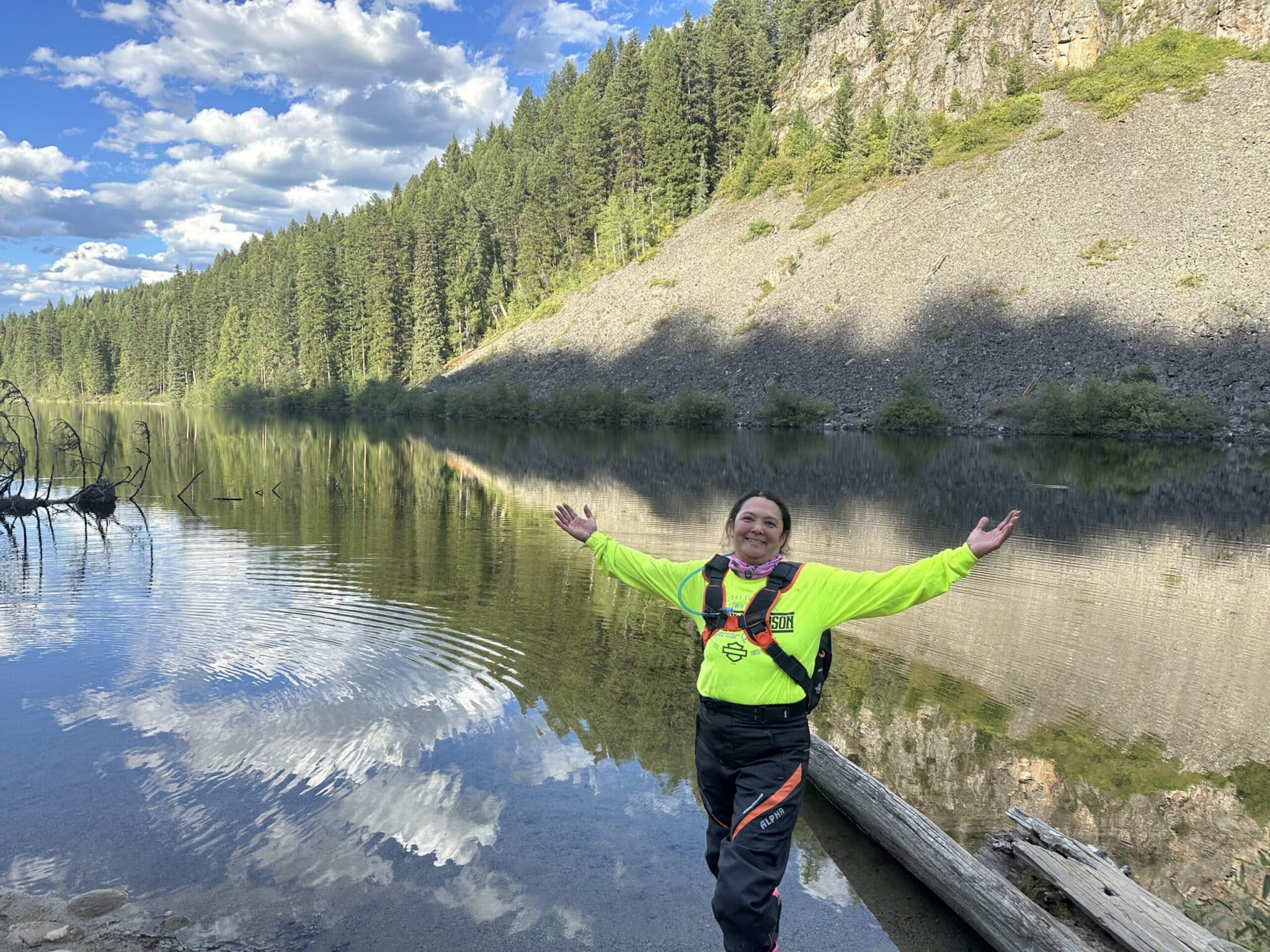 Woman in bright green shirt standing by a reflective mountain lake with forest and rocky hillside background under a partly cloudy sky.