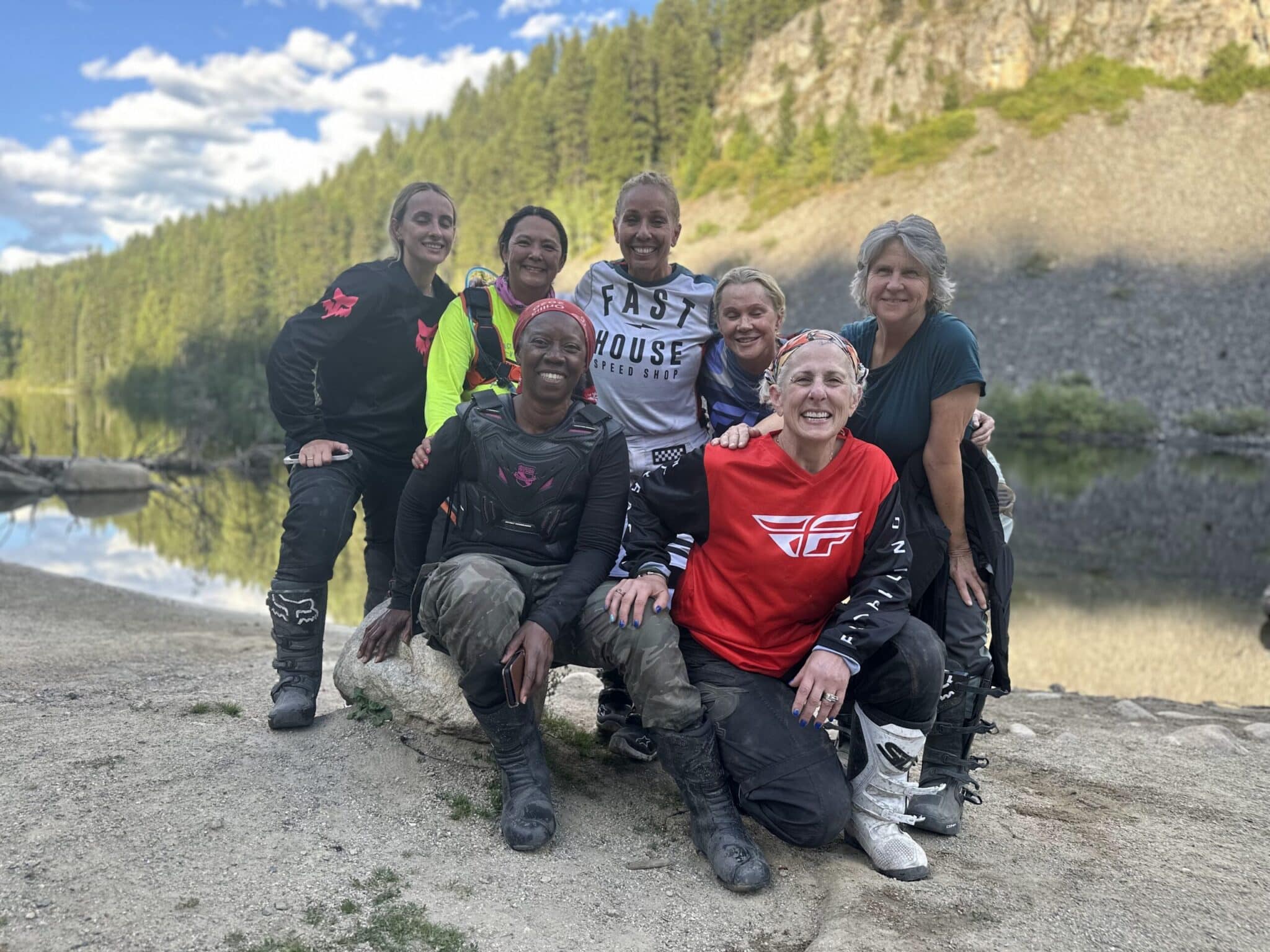 Group of seven women in motocross gear posing outdoors near a lake with forest and rocky hills in the background.