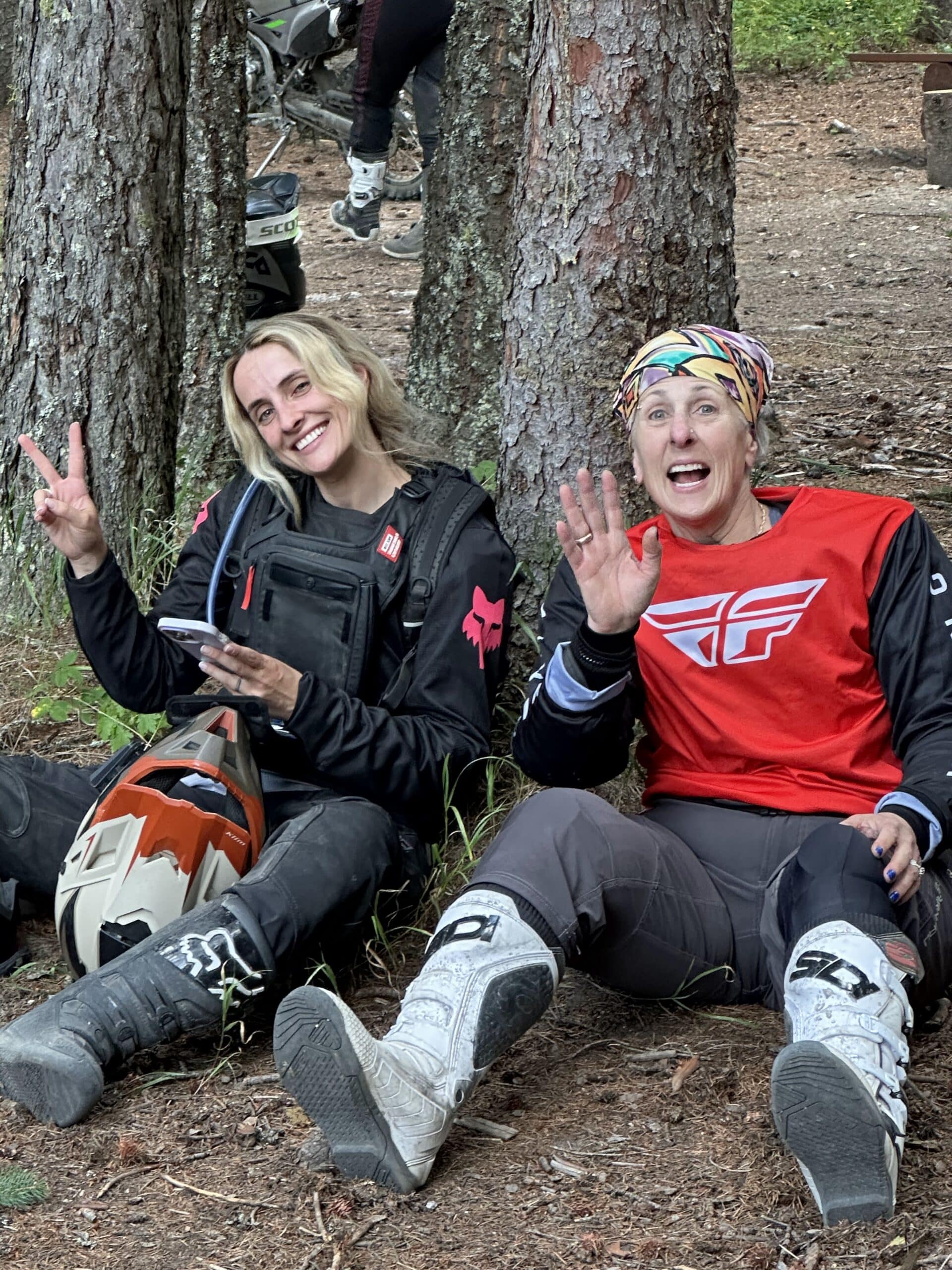 Two women in motocross gear sitting on forest ground by trees, one holding a helmet and phone, both smiling and waving.