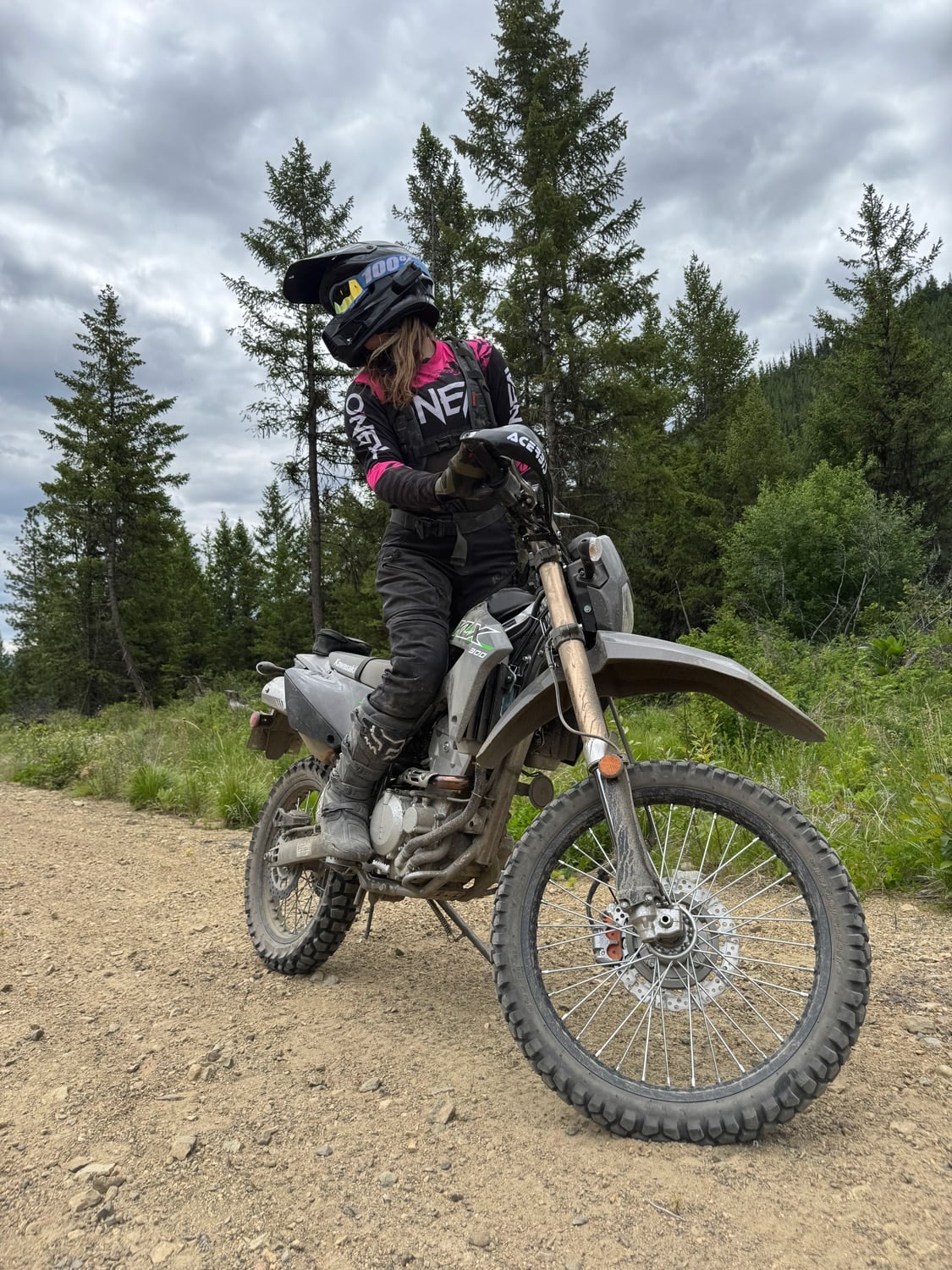 Woman wearing full motocross gear riding a dirt bike on a forest trail under cloudy sky.