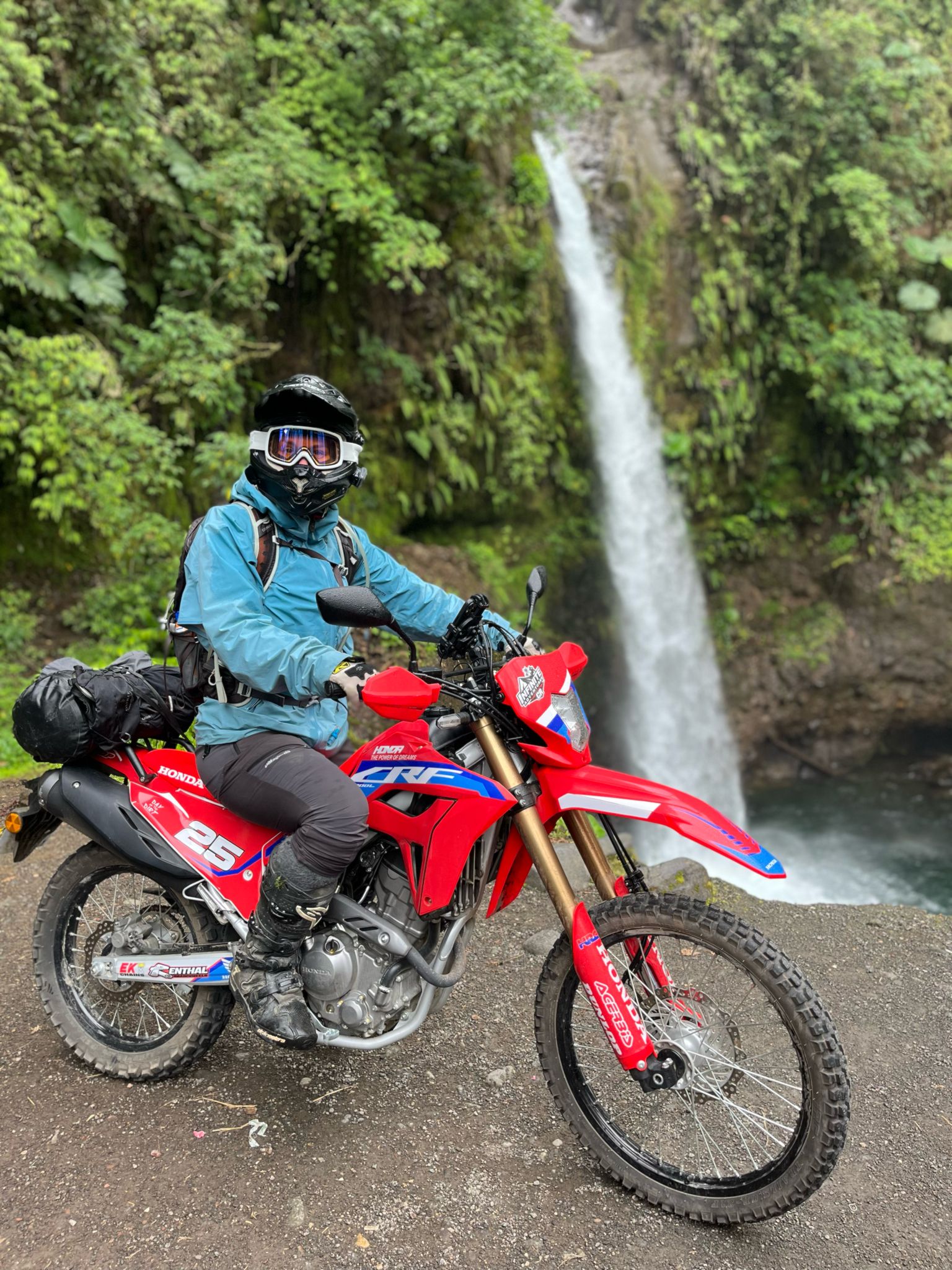 Motorcyclist in protective gear on red Honda dirt bike near a forest waterfall.