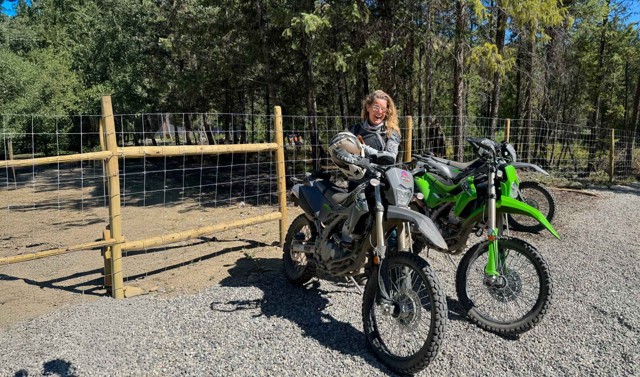 Person standing next to two dirt bikes, a gray model and a green model, in a gravel area surrounded by trees and a wooden fence.