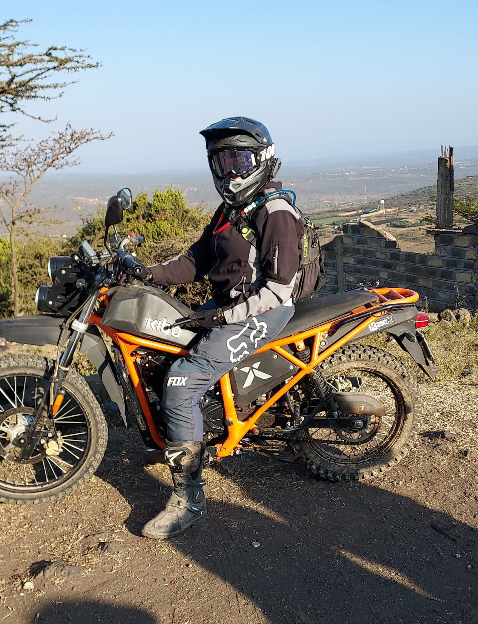 Person wearing protective gear and a helmet, sitting on an orange off-road motorcycle in a scenic outdoor setting.