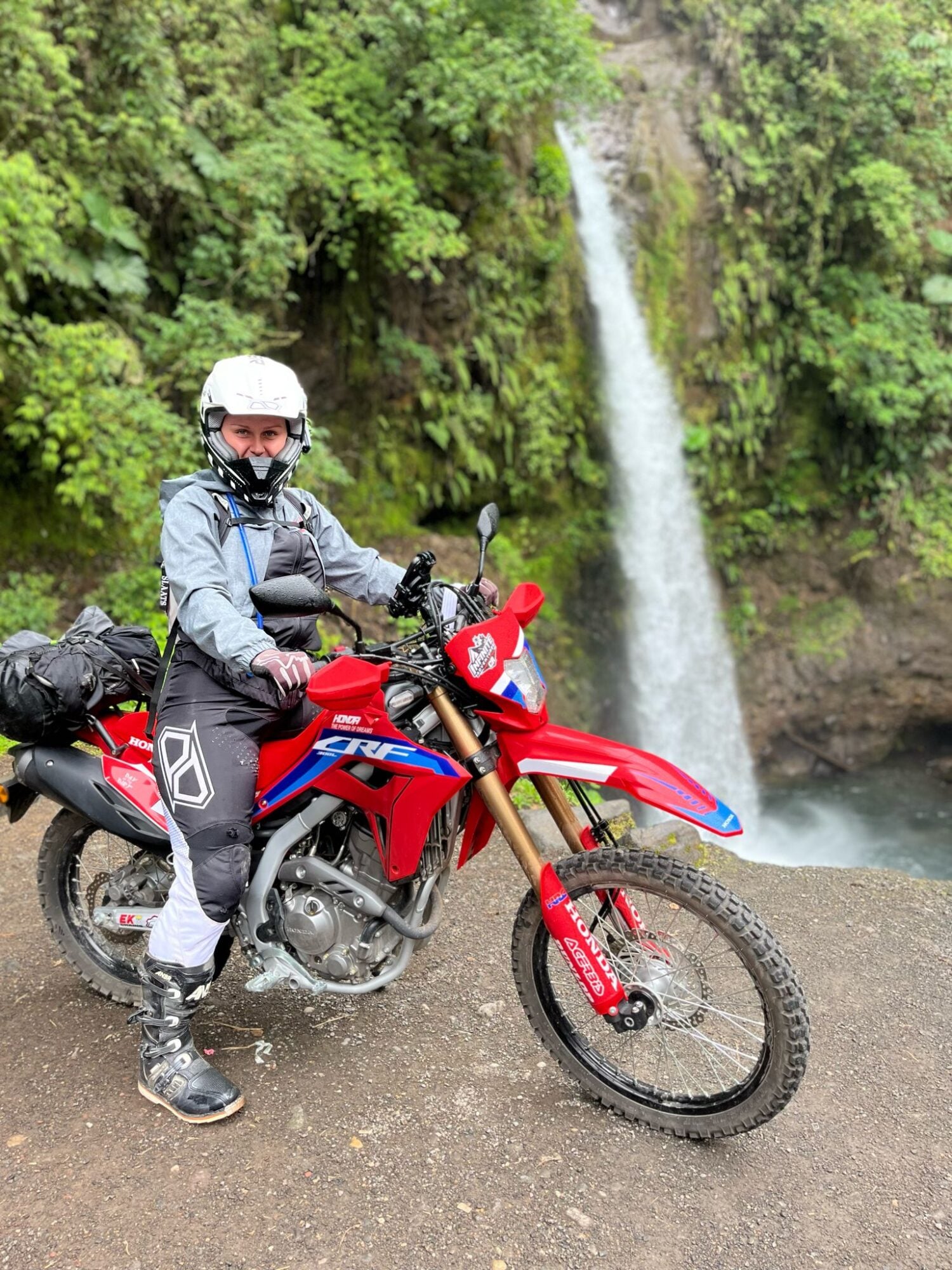 Woman wearing protective gear sitting on red Honda dirt bike near waterfall in lush green forest.