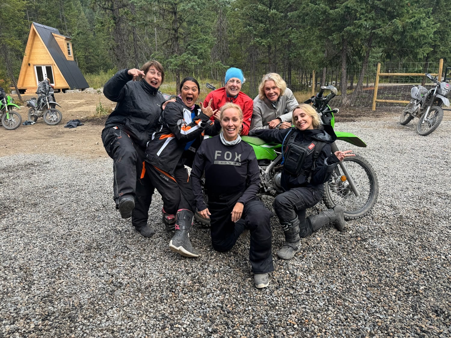 Group of six women in motorcycle gear posing outdoors near dirt bikes and a wooden cabin in a forest setting.
