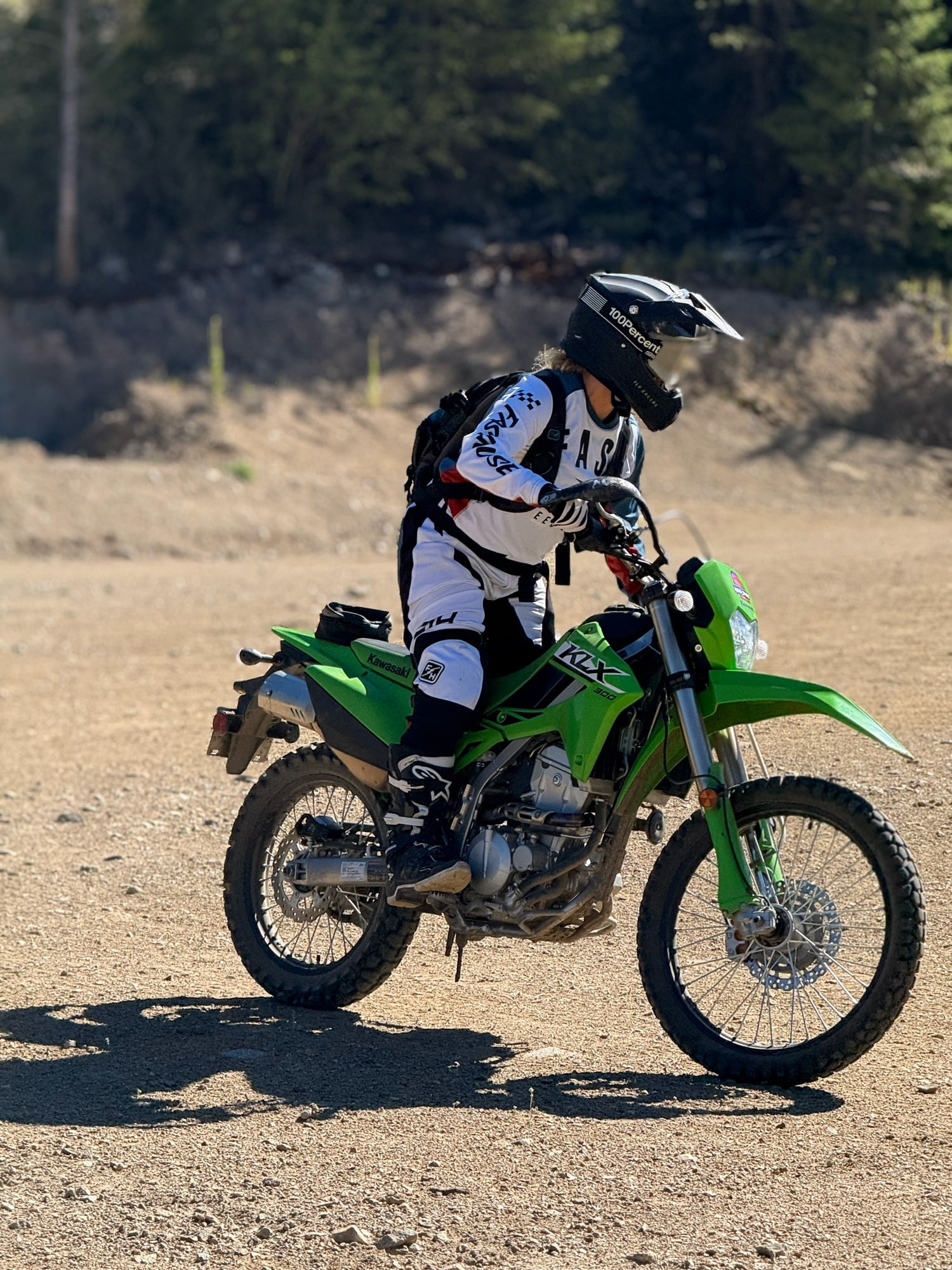 Woman wearing motocross gear and helmet sitting on a green Kawasaki KLX 300 dirt bike on a dirt trail.