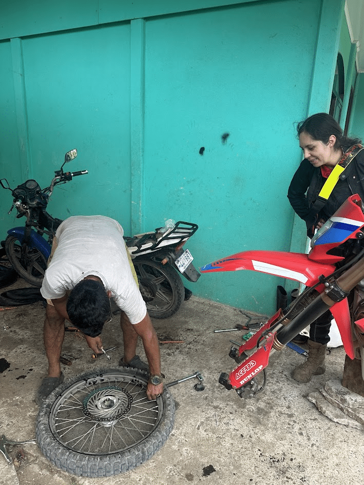 Man repairing motorcycle tire while woman watches in a workshop with multiple motorcycles and tools.