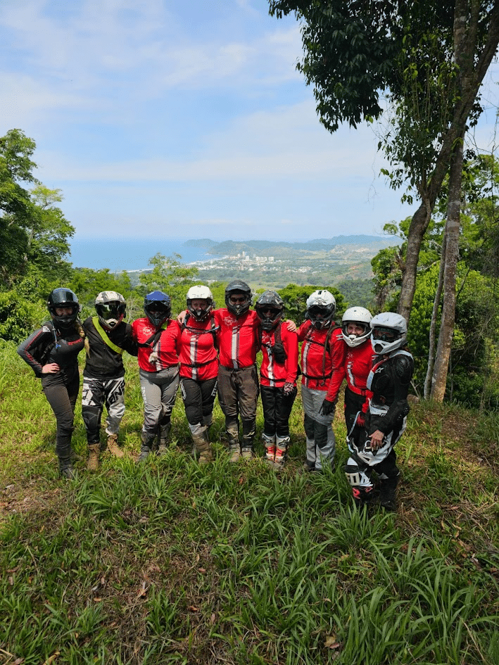 Group of motorcyclists in red jackets posing for a photo on a scenic hillside, with a view of the ocean and lush greenery in the background.