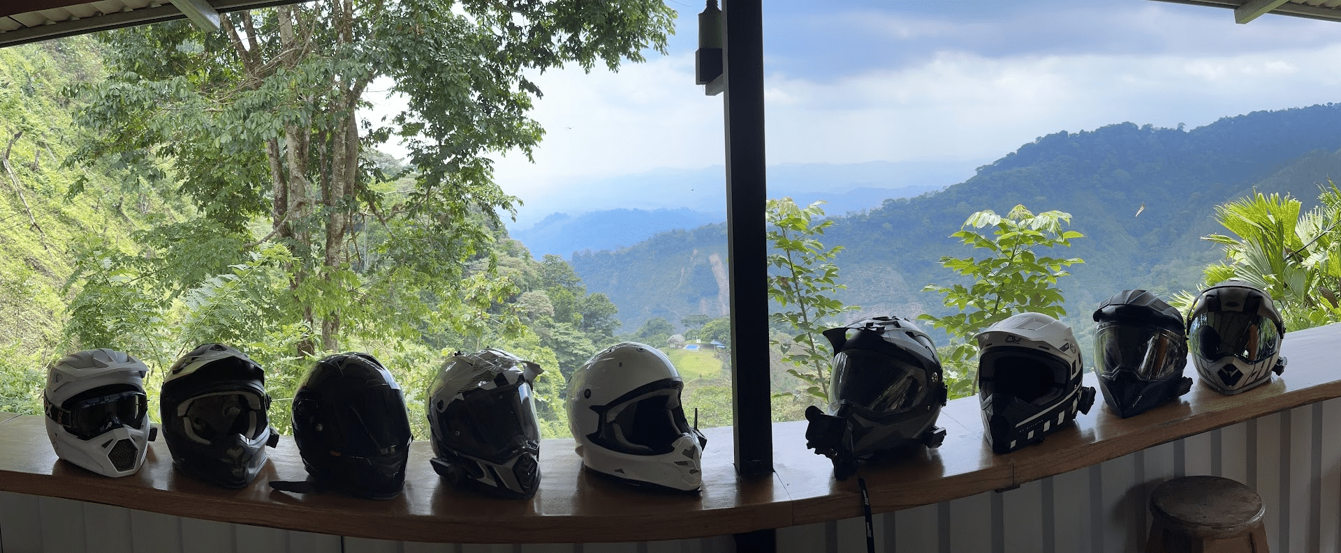 Four motorcycle helmets displayed on a wooden table with a scenic mountain view in the background, featuring lush greenery and a cloudy sky.