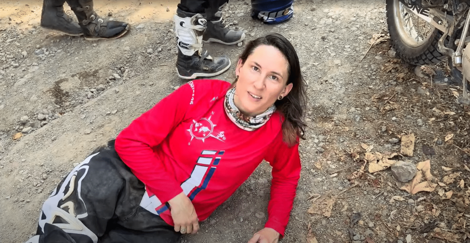 A person wearing a red athletic shirt and a neck gaiter is sitting on the ground in an outdoor setting, surrounded by other people in hiking gear.