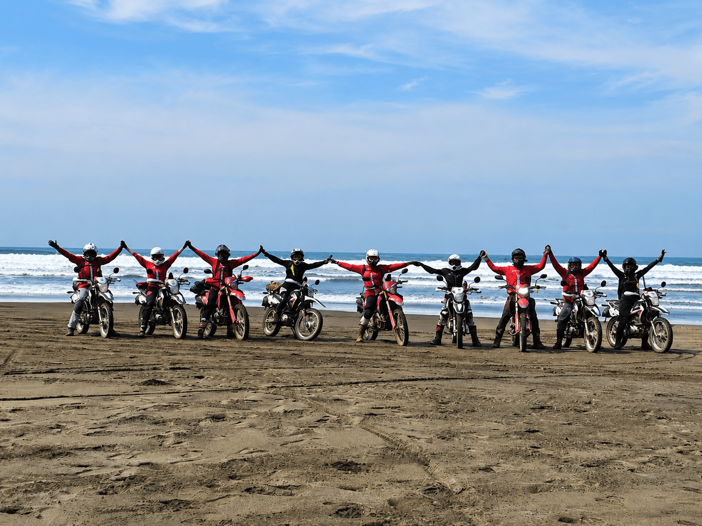 Group of motocross riders on a beach, standing beside their bikes with arms raised, enjoying a sunny day by the ocean.