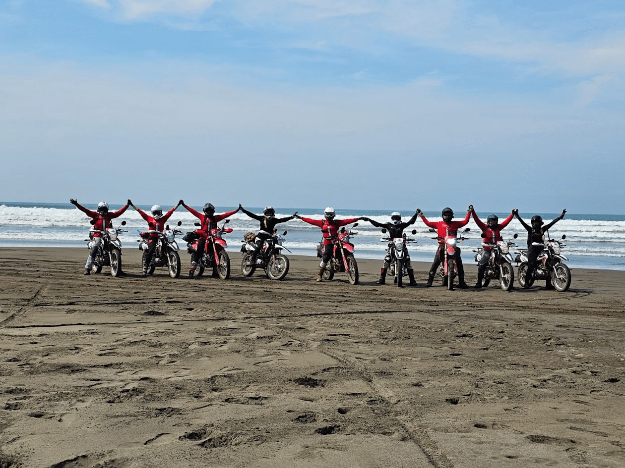 Group of motocross riders on a beach, standing beside their bikes with arms raised, enjoying a sunny day by the ocean.