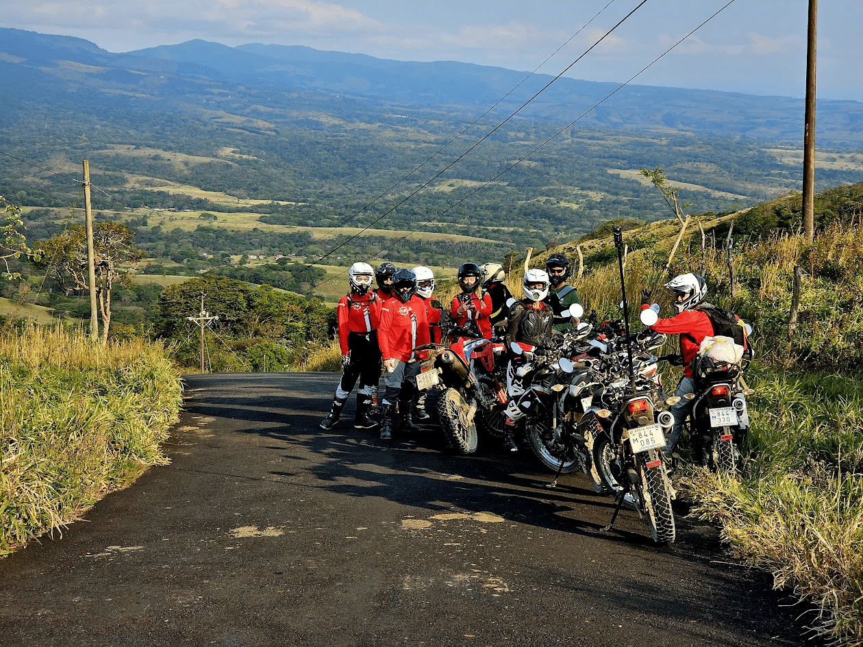 Group of motorcyclists in red jackets parked on a scenic mountain road, overlooking a lush valley and distant mountains, ideal for adventure and outdoor enthusiasts.