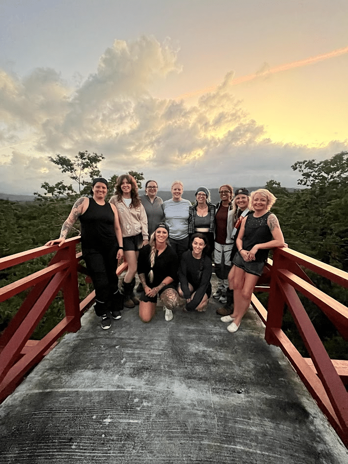 Group of women posing on a red wooden bridge at sunset with a scenic forest background.
