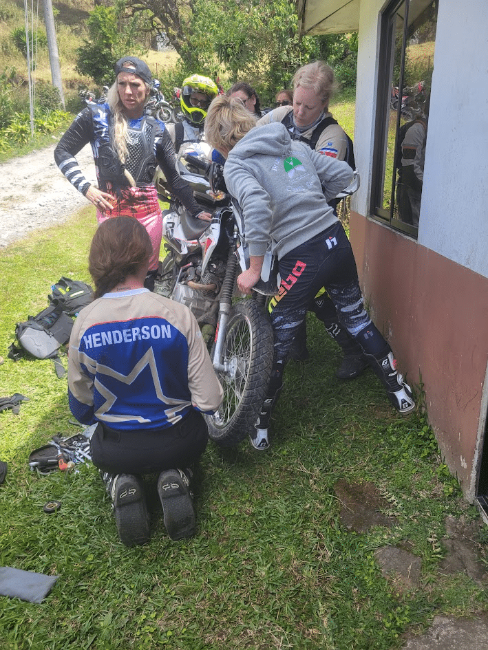 Group of motocross riders working together to repair a motorcycle outdoors, showcasing teamwork and mechanical skills in a grassy area.