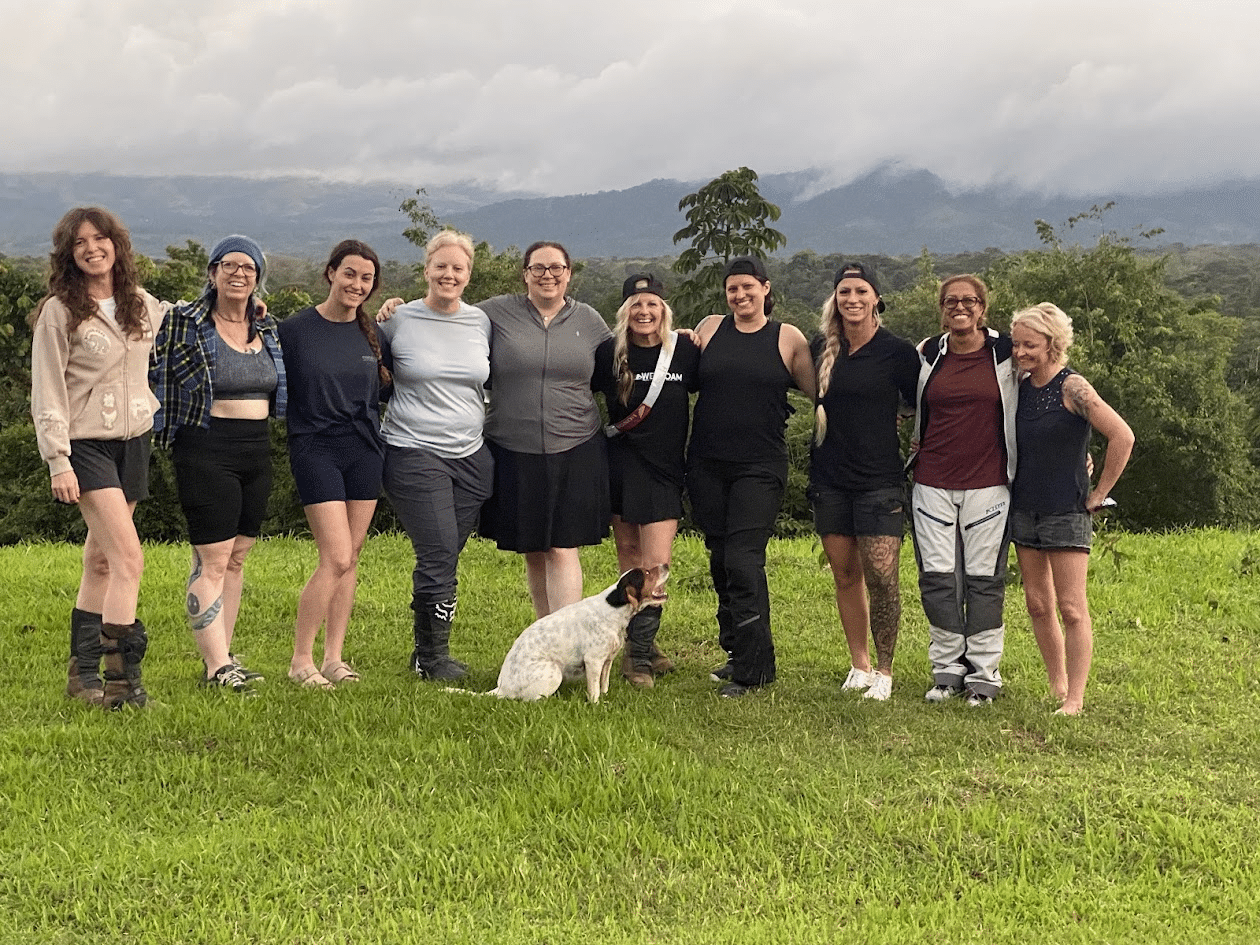 Group of women standing together outdoors on a grassy field with mountains in the background, accompanied by a dog. The image captures a moment of camaraderie and friendship.