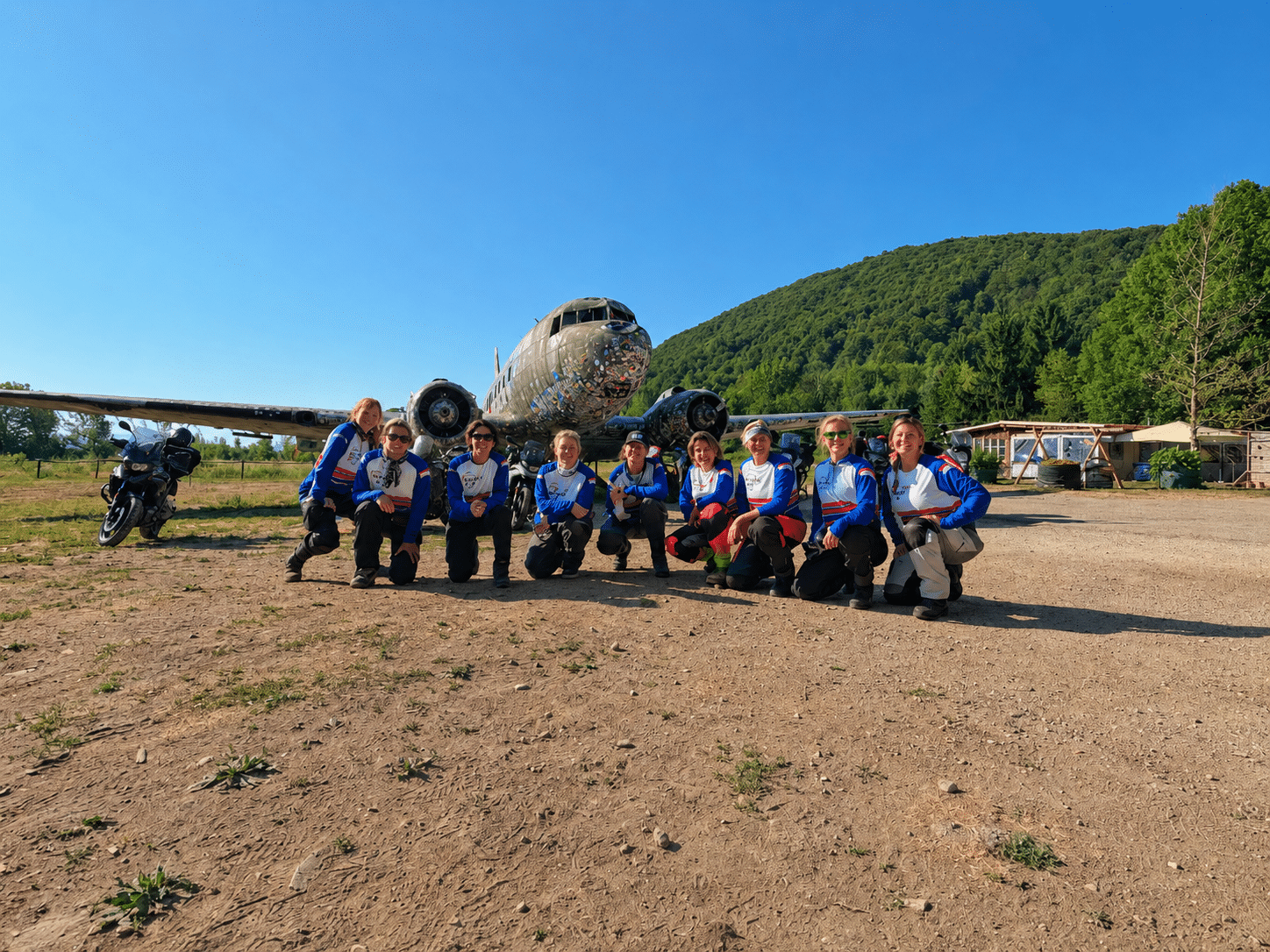 Group of women in matching blue and white shirts posing in front of a vintage airplane with a green hill and clear blue sky in the background.