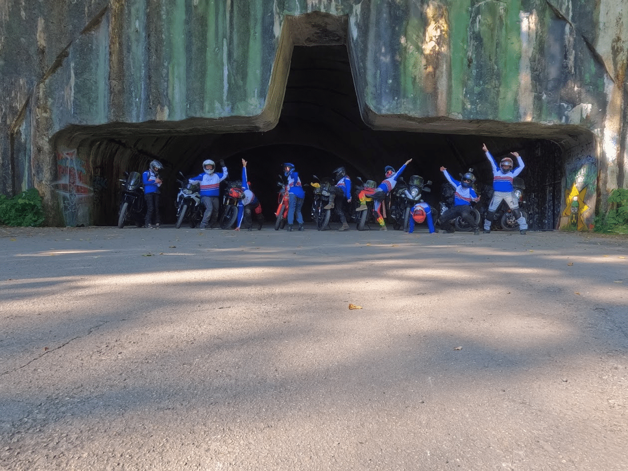 Group of motorcyclists posing and doing handstands in front of a tunnel shaped like a cross with graffiti on the walls.