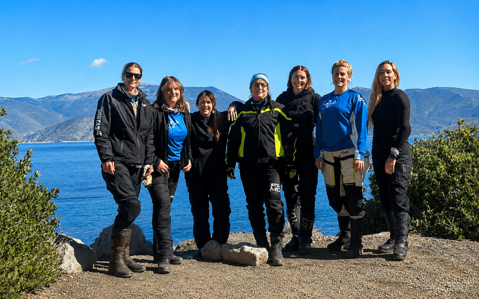 Group of seven women in outdoor gear standing on a rocky shoreline with a scenic ocean view and mountains in the background, promoting adventure and camaraderie in outdoor activities.