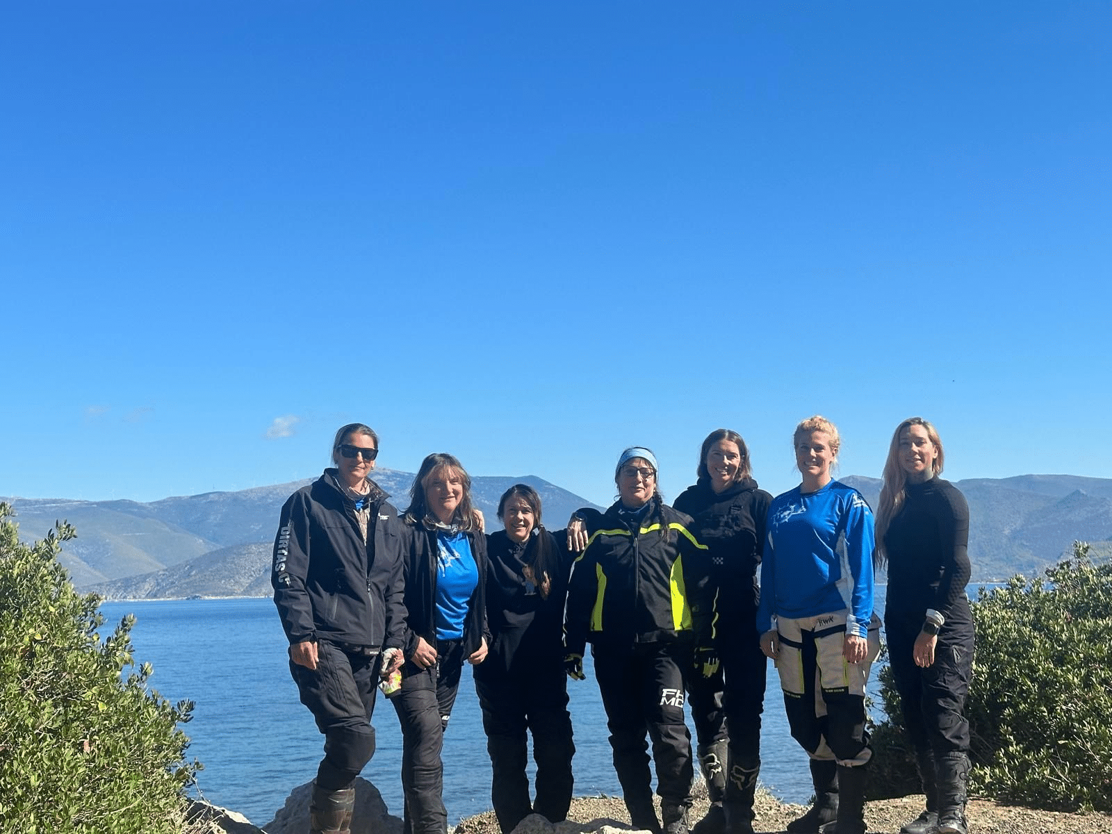 Group of six people standing together by the ocean, wearing outdoor gear and smiling, with mountains and a clear blue sky in the background.