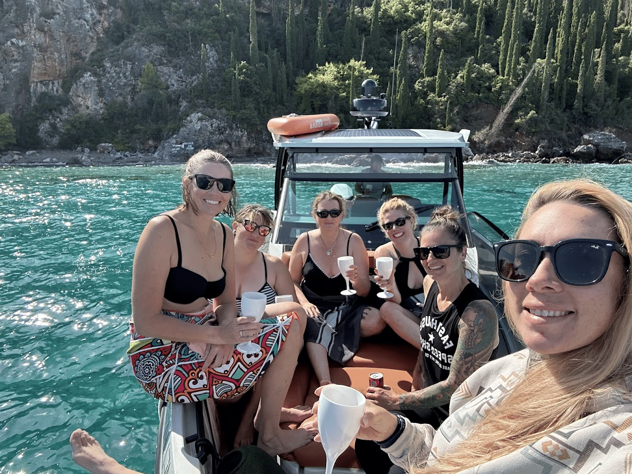 Group of women enjoying a sunny day on a boat, smiling and holding drinks, with a scenic backdrop of lush greenery and clear blue water.