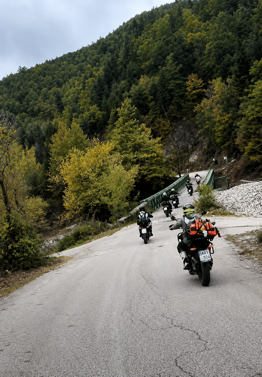 Group of motorcyclists riding on a winding mountain road through dense green forest with a small bridge ahead.
