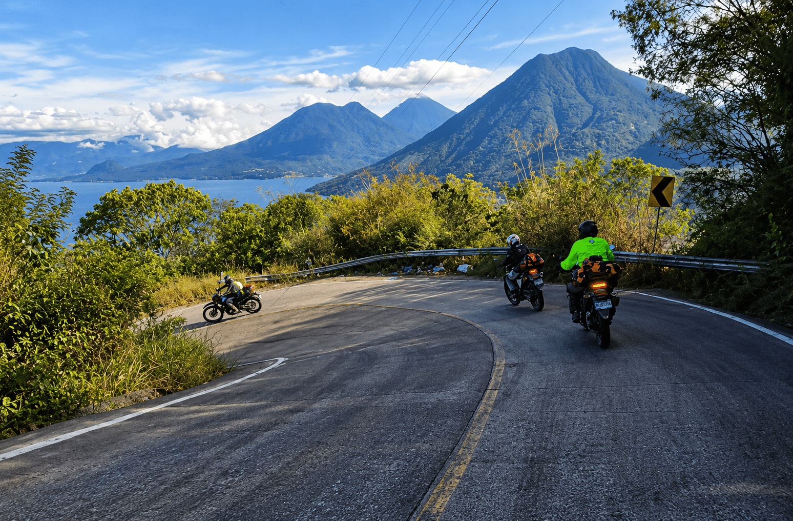 Motorcyclists riding on a winding mountain road with lush greenery and volcanic mountains in the background under a blue sky.