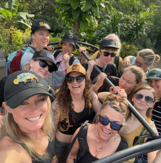 Group of smiling women wearing hats and sunglasses outdoors, surrounded by greenery.