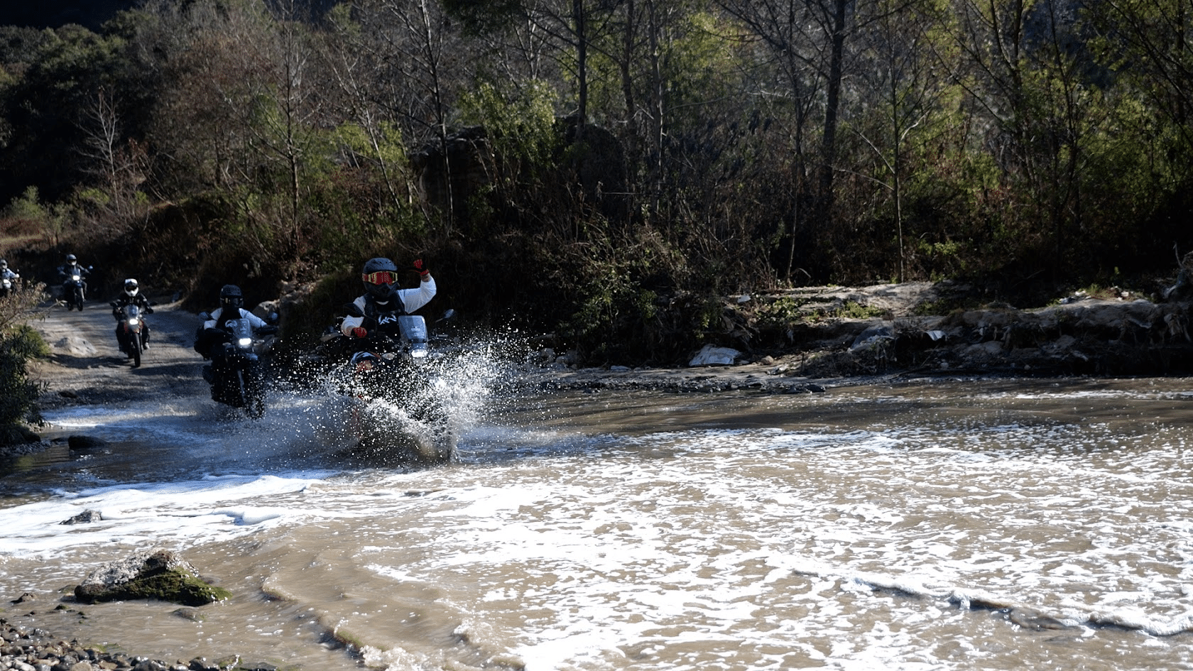 Motorcyclist riding through a shallow river, splashing water, surrounded by lush greenery and trees in a natural outdoor setting.