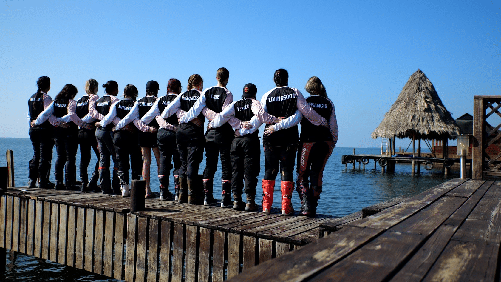 Group of people standing on a wooden dock by the water, wearing matching jackets, with their backs to the camera, enjoying a sunny day outdoors.