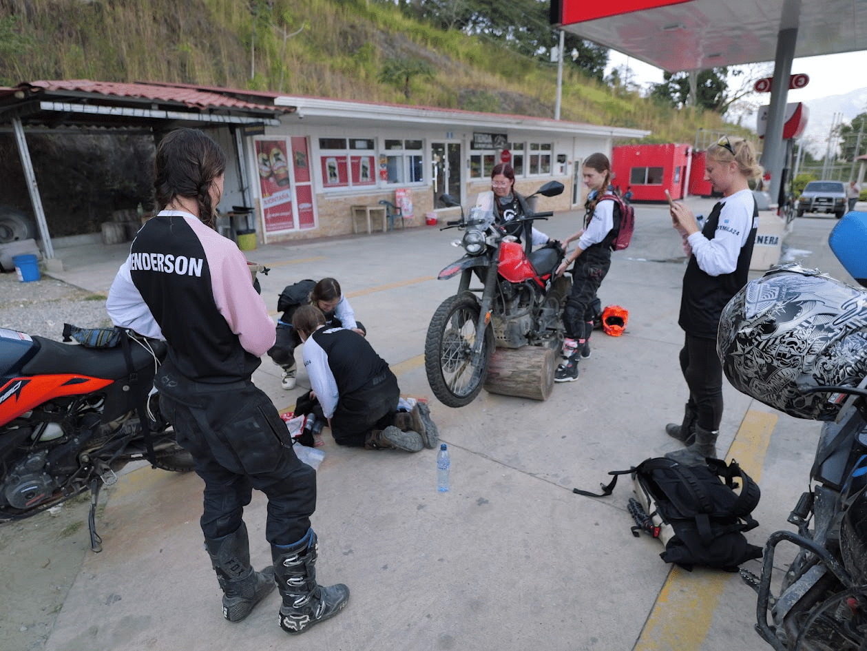 Group of motorcyclists in riding gear gathered at a gas station, with one person working on a motorcycle and others observing. The setting features a rural backdrop and various motorcycle gear scattered around.
