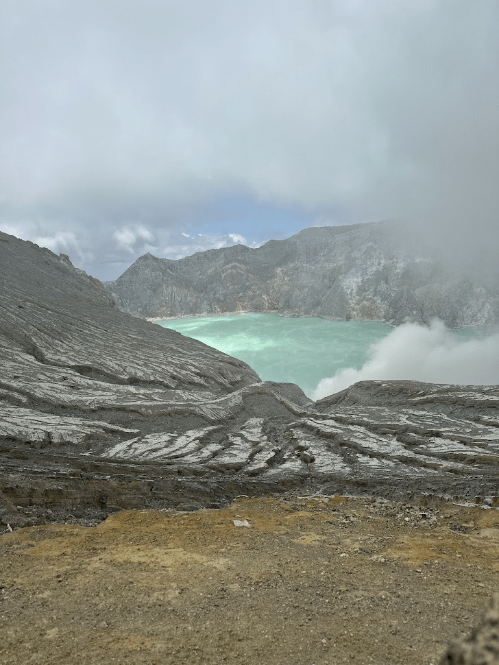 Stunning panoramic view of a volcanic crater lake surrounded by rugged terrain and misty clouds, showcasing the vibrant turquoise water and volcanic rock formations.