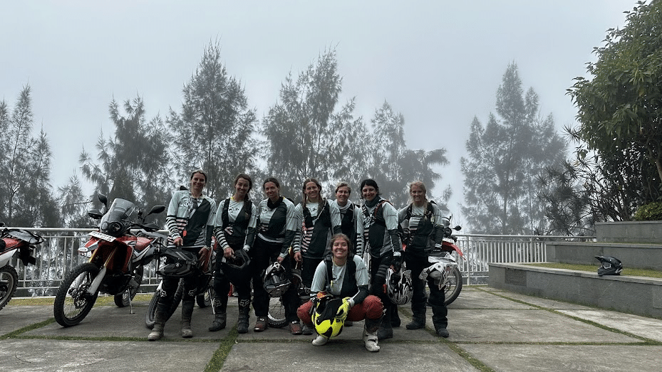 Group of female motorcyclists posing together with their bikes in a foggy outdoor setting, showcasing camaraderie and adventure in a scenic location.