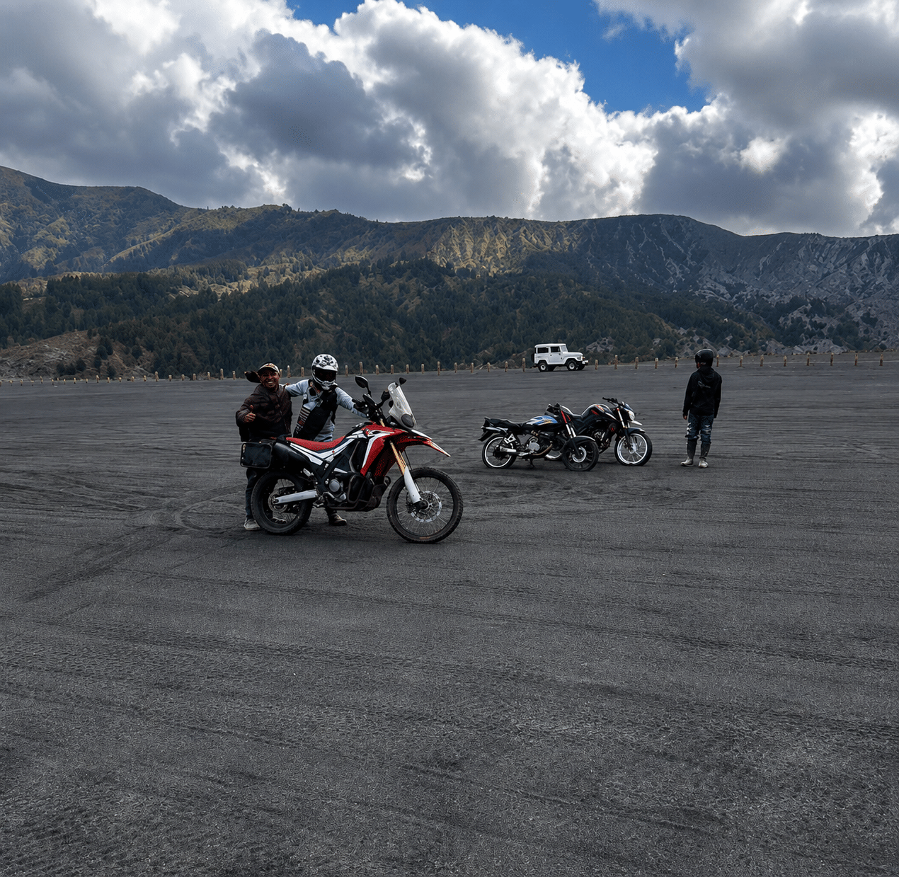 Two motorcyclists on an adventure in a vast, open terrain with mountains in the background, showcasing off-road motorcycles and a cloudy sky.