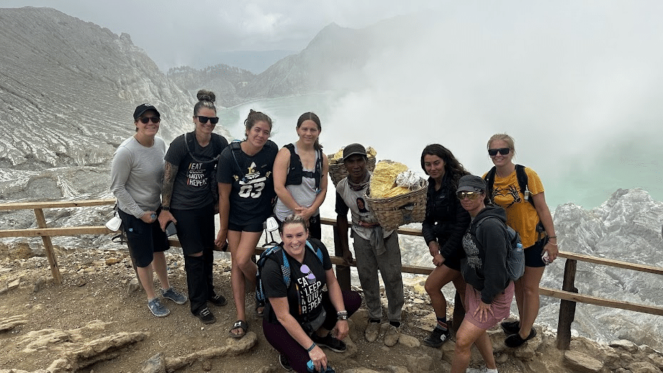 Group of hikers posing at a mountain summit with a local guide, surrounded by misty peaks and scenic views. The group is diverse, wearing hiking gear, and some are carrying backpacks.