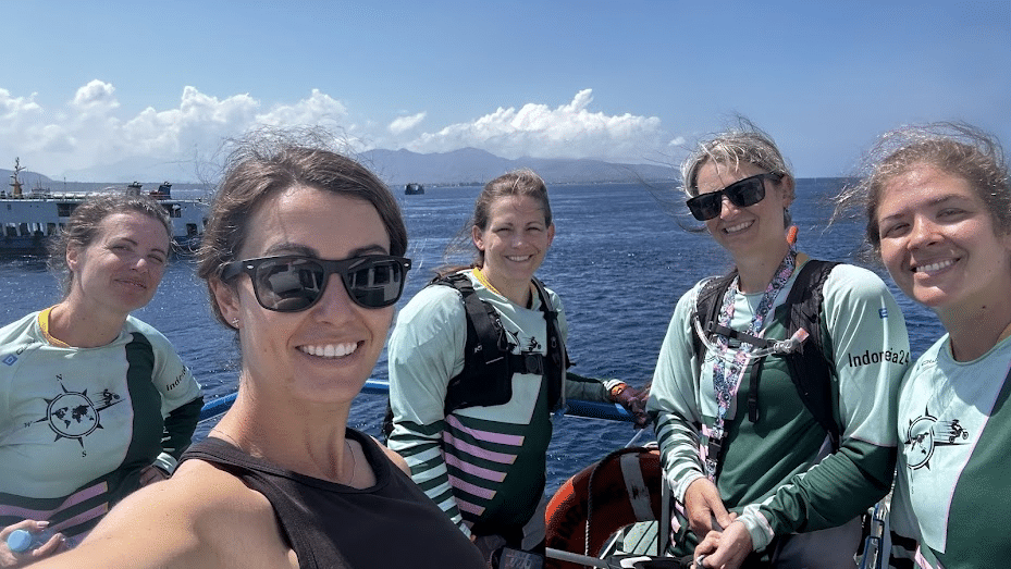 Group of five women smiling on a boat with ocean and mountains in the background on a sunny day.